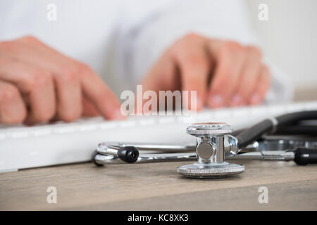 Nahaufnahme der männlichen Arzt mittels Computer Tastatur mit Stethoskop auf hölzernen Schreibtisch in der Klinik Stockfoto