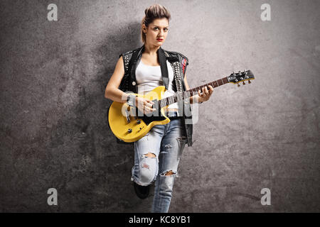 Weibliche Punker spielen eine E-Gitarre und lehnte sich gegen eine rostige graue Wand Stockfoto