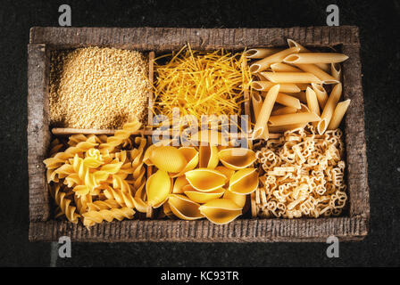 Auswahl von verschiedenen Rohstoffen italienische Pasta mit Basilikum und Cherry Tomaten, in alten hölzernen Kasten auf dunklem Stein Tabelle, Ansicht von oben kopieren Raum Stockfoto
