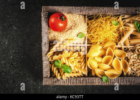 Auswahl von verschiedenen Rohstoffen italienische Pasta mit Basilikum und Cherry Tomaten, in alten hölzernen Kasten auf dunklem Stein Tabelle, Ansicht von oben kopieren Raum Stockfoto