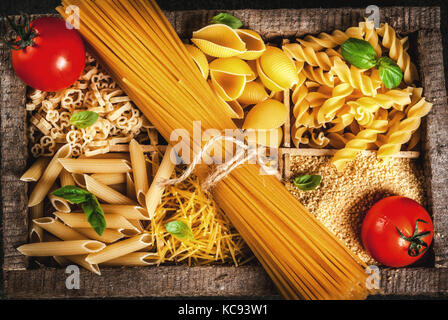 Auswahl von verschiedenen Rohstoffen italienische Pasta mit Basilikum und Cherry Tomaten, in alten hölzernen Kasten auf dunklem Stein Tabelle, Ansicht von oben kopieren Raum Stockfoto