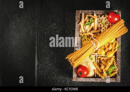 Auswahl von verschiedenen Rohstoffen italienische Pasta mit Basilikum und Cherry Tomaten, in alten hölzernen Kasten auf dunklem Stein Tabelle, Ansicht von oben kopieren Raum Stockfoto