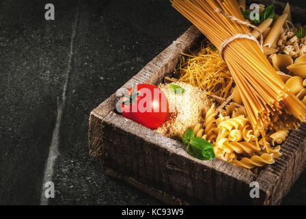 Auswahl von verschiedenen Rohstoffen italienische Pasta mit Basilikum und Cherry Tomaten, in alten hölzernen Kasten auf dunklem Stein Tabelle, Ansicht von oben kopieren Raum Stockfoto
