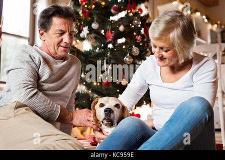 Senior Paar sitzt auf dem Boden vor der beleuchteten Weihnachtsbaum in ihrem Haus mit ihren Hund. Stockfoto