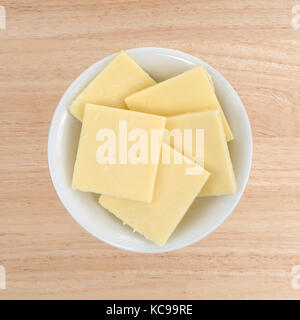 Blick von oben auf einem kleinen weißen Schale mit Sharp Cheddar Käse quadrat Scheiben auf einem Holz Tisch gefüllt. Stockfoto