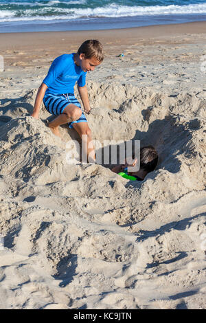 Avon, Outer Banks, North Carolina, USA. Junge Junge die Kontrolle über seinen Bruder, begraben in den Sand. Stockfoto