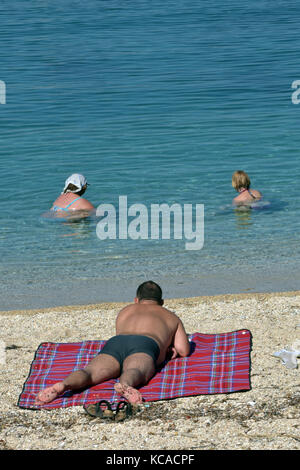 Eine große gebräunte Mann mittleren Alters mit schwarzen Badehose lag auf einem Strand bei zwei Frauen schwimmen im Meer vor ihm. Sonnenbaden. Stockfoto