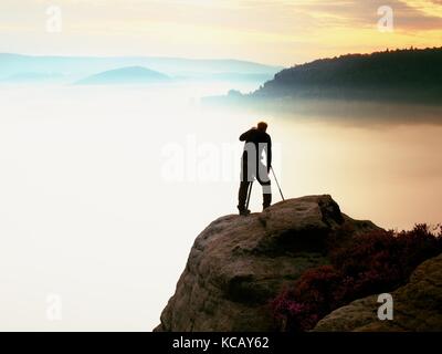 Professioneller Fotograf nimmt Fotos mit Kamera und Stativ auf der Spitze des Felsens. verträumte fogy Landschaft, orange rosa Nebel Sonnenaufgang in beaut Stockfoto