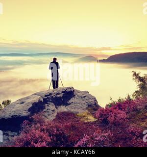 Professioneller Fotograf nimmt Fotos mit Kamera und Stativ auf der Spitze des Felsens. verträumte fogy Landschaft, orange rosa Nebel Sonnenaufgang in beaut Stockfoto