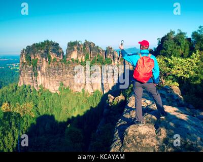 Touristische mit roten Rucksack nimmt Fotos mit Smart Phone von Felsen und Tal. im Frühling oder im Sommer bei schönem Wetter, frische grüne Farben Stockfoto