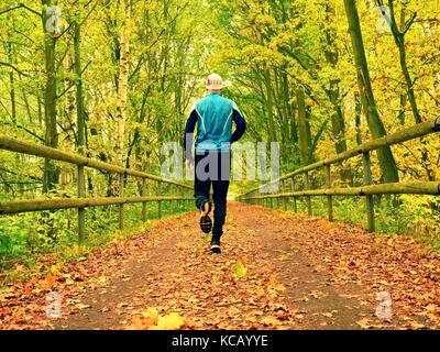 Sportsman in blauen T-Shirt und schwarze Hose laufen auf der Straße. Der Mann wird langsam laufen auf Asphalt Art und Weise durch die Blätter im Herbst fallen. Bahn im Park, beeches Stockfoto