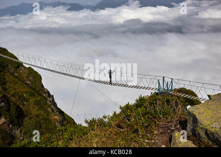 Man Walking auf Suspension Bridge und bei bewölktem Berge unterhalb der Suche Stockfoto