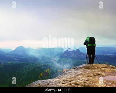 Professionelle Fotografen in grüne Jacke mit Rucksack Fotos mit Kamera auf Stativ auf felsigen Gipfel. verträumte fogy Landschaft, schöne Tal bel Stockfoto