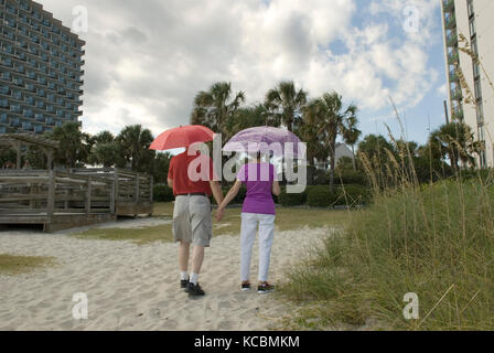 Kaukasisches Senior-Paar (60-70) mit Sonnenschirmen beim Spazierengehen auf dem Pfad, Myrtle Beach SC, USA. Stockfoto