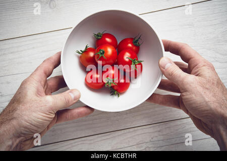 A bemannt Händen herauf eine weiße Schale, leuchtend rote reife Tomaten auf einer Arbeitsfläche aus hellem Holz, Arbeitsplatte in der Küche. Stockfoto