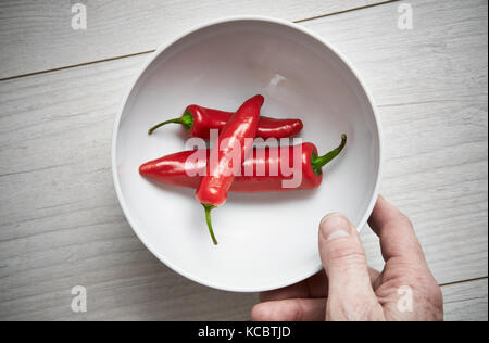 A bemannt Händen herauf eine weiße Schale, helle rote reife Chilischoten auf hellem Holz Arbeitsfläche, Arbeitsplatte in der Küche. Stockfoto