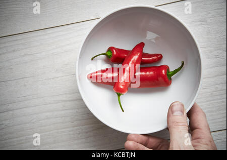 A bemannt Händen herauf eine weiße Schale, helle rote reife Chilischoten auf hellem Holz Arbeitsfläche, Arbeitsplatte in der Küche. Stockfoto