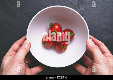 Mans Händen herauf ein weißer Keramik Schüssel mit leuchtend rote reife Tomaten auf einem dunkelgrauen Schiefer Arbeitsfläche, Arbeitsplatte in der Küche. Stockfoto