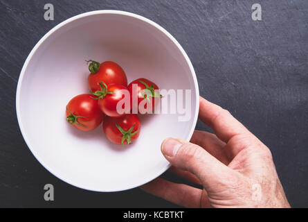 Mans Händen herauf ein weißer Keramik Schüssel mit leuchtend rote reife Tomaten auf einem dunkelgrauen Schiefer Arbeitsfläche, Arbeitsplatte in der Küche. Stockfoto