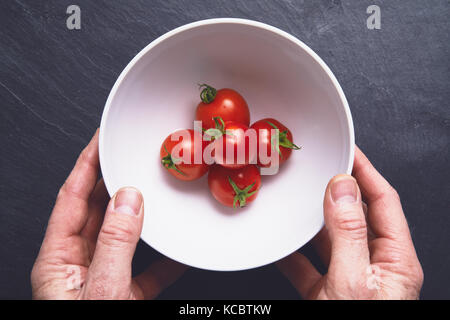 Mans Händen herauf ein weißer Keramik Schüssel mit leuchtend rote reife Tomaten auf einem dunkelgrauen Schiefer Arbeitsfläche, Arbeitsplatte in der Küche. Stockfoto