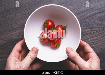 Mans Händen herauf ein weißer Keramik Schüssel mit leuchtend rote reife Tomaten auf einem dunklen Holz Arbeitsfläche, Arbeitsplatte in der Küche. Stockfoto