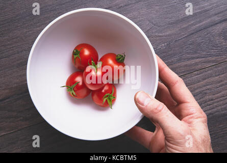 Mans Händen herauf ein weißer Keramik Schüssel mit leuchtend rote reife Tomaten auf einem dunklen Holz Arbeitsfläche, Arbeitsplatte in der Küche. Stockfoto