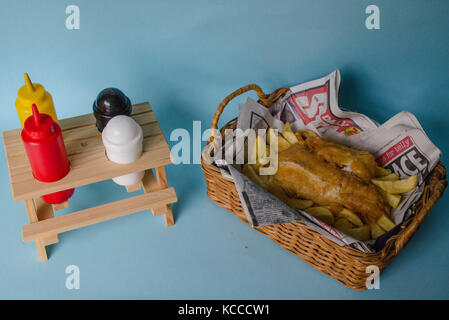 Die traditionelle Fish n Chips serviert in einem Korb mit Zeitung. Pick Nick Bank Tabelle Würzmittel Fach auf blauem Hintergrund. Stockfoto