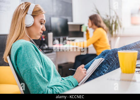 Junge geschäftsfrau am Schreibtisch mit ihren Beinen auf dem Tisch sitzen und Musik hören mit Kopfhörern auf dem Kopf und Notizen Stockfoto