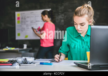 Young Business Frau trägt Grün shirt Schreibarbeit zu tun im Büro und im Hintergrund ist Frau Schreiben auf dem Whiteboard Stockfoto