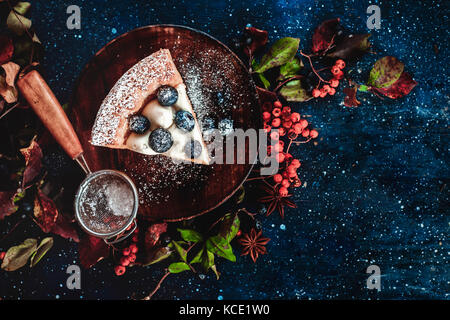 Close-up ein Stück Pumpkin Pie mit Puderzucker und ein Sieb auf einer hölzernen Platten. handgefertigte Gerichte im Herbst flach. Konzeptionelle stilisierte Essen noch leben, Table Top Shot auf dunklem Hintergrund. Stockfoto