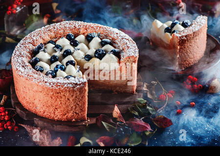 Cut Kuchen mit einem Shortbread Kruste auf dunklem Hintergrund. Ein Stück Kuchen mit Schlagsahne und Heidelbeeren auf einer Holzplatte. Dunkle essen Fotografie mit Rauch. Kontrast Farben und weichen konzentrieren. Stockfoto