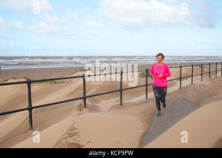 Frau beim Joggen in Crosby, Merseyside. Wetter in Großbritannien. Oktober 2017. Starke Winde wehen feinen leichten Sand vor dem Strand, während Stürme weiterhin die Westküste und Mersey Mündung schlagen. Dünenbau Akkretion wird entlang durch die charakteristischen Onshore Küstenwinde geholfen. Bulldozing hat sich zu einer gemeinsamen Form der künstlichen Dünenkonstruktion, zum Teil, weil die Vegetation & Fechten Ansatz dauert einige Zeit, um Sand zu fangen und eine neue Düne zu bauen. Stockfoto