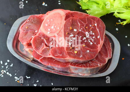 Rohes Rindfleisch Fleisch mit Knochen. ossobuco Fleisch auf einem Metallauflagefach auf einem grauen Abstrakt Hintergrund mit einigen grünen frischen Sellerie verlässt. traditionellen italienischen Essen Stockfoto