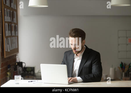 Junger gutaussehender Geschäftsmann im Anzug, der am Laptop am Schreibtisch in einem modernen Büro arbeitet. Der CEO kümmert sich um wichtige Aufgaben, schreibt E-Mails, leitet den Projektmanager Stockfoto