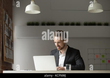 Junger, fröhlicher Geschäftsmann, der im modernen Büro sitzt, mit einem glücklichen Lächeln auf den Laptop blickt, zufrieden mit dem erfolgreichen Abschluss, dem Finanzbericht, dem Projektresu Stockfoto