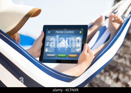 Verwendung von Remote Home Control System, das auf digitaler Tablet während auf Hängematte am Strand liegende Frau Stockfoto
