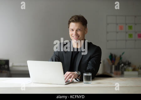 Portrait von Stattlichen lächelnd Geschäftsmann in Anzug im modernen Büro vor dem Laptop, an der Kamera schaut. positive Person während der wichtigen Mee Stockfoto