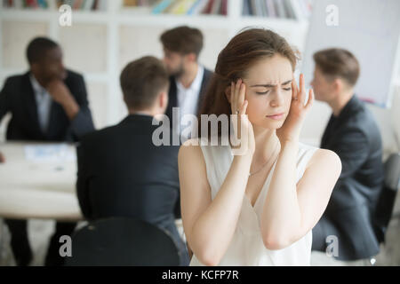 Gestresste junge Unternehmensangestellte mit Migräne bei unproduktivem Briefing. Geschäftsfrau bekam Kopfschmerzen von stressigem Meeting, Kollegen nicht c Stockfoto