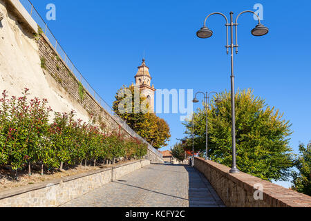 Schmale Straße, die in Richtung Glockenturm entlang Strassenlaternen und herbstliche Bäume unter blauem Himmel in der kleinen Stadt von Diano d'Alba in Piemont, Norditalien. Stockfoto