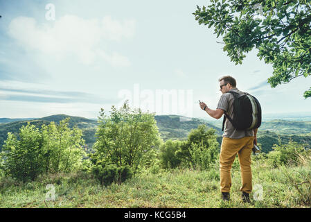 Mann mit Sonnenbrille und Rucksack ruhen unter dem Baum mit Wasserflasche und Kontrolle smart phone Stockfoto