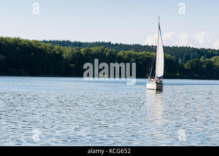 Kleine Yacht segeln am Lipno-See. Wald auf Hintergrund, Reflexion über Wasser und Wolken am Himmel. helle Sommer sonnigen Tag. Stockfoto