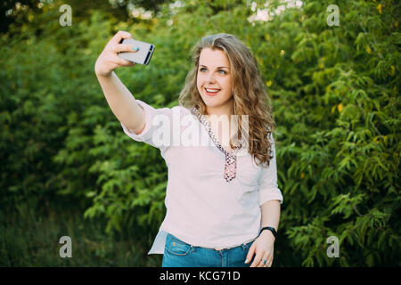 Junge schöne ziemlich Plus size kaukasische Mädchen Frau in weißen Bluse das Leben genießen gekleidet, lächelnd, Spaß und unter selfie Foto auf dem Smartphone Stockfoto