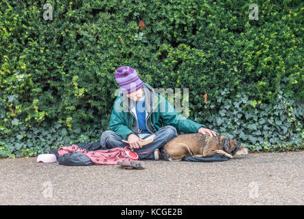 Ältere/alte/Ältere obdachlose Bettler Mann/rough Sleeper, das Tragen einer Mütze Hut, sitzen, ein Buch lesen, mit einer Hand auf seinen Hund, London, England, UK. Stockfoto