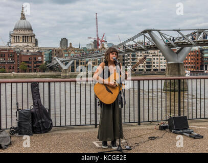 Eine junge Frau, die Gaukler, singen und spielen Gitarre, mit Mikrofon und tragbaren Verstärker, neben der Millennium Bridge, London, England, UK. Stockfoto