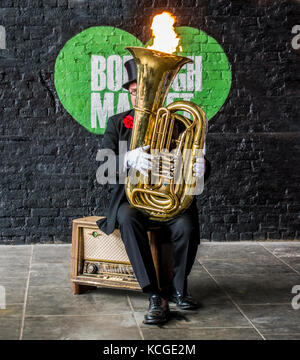 Man Entertainer in Anzug und Hut, sitzend auf einem alten Grammophon, spielen eine Tuba mit einer Flamme von oben. Borough Market, London, England, Großbritannien Stockfoto