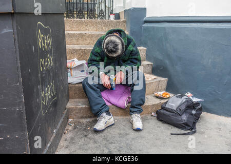 Ein Betrunkener, obdachlosen Mann sitzt auf Schritte, die eine Dose Bier, mit einem halben Mahlzeit in der Nähe gegessen, mit dem Kopf nach unten und mit einem Hut bedeckt. London, England, Großbritannien Stockfoto