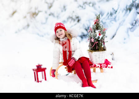 Kinder mit Weihnachtsbaum auf hölzernen Schlitten im Schnee. Kinder schneiden Xmas Tree. Kleines Mädchen auf Schlitten im verschneiten Wald. Familie auswählen Winterurlaub Dekor Stockfoto