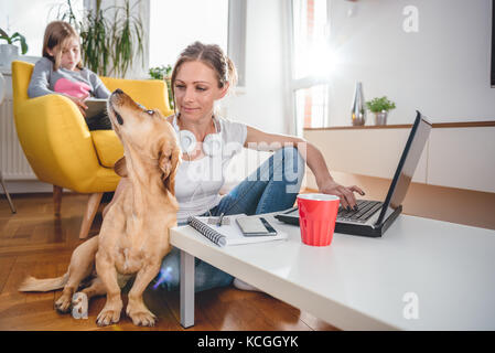 Frau sitzt am Tisch auf den Boden und streichelte Hund zu Hause, während die Tochter sitzt auf dem Sessel und mit Tablet Stockfoto