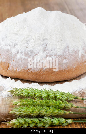 Ein Stück Roggenbrot und grüne, unreife Roggenohren liegen auf einem Holztisch. Hausgemachte Küche. Nahaufnahme von Fotos. Weißmehl wird über das Brot gegossen Stockfoto