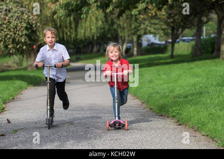 Kinder reiten ihre Roller - ein kleiner Junge und seine kleine Schwester, die Spaß Rennen entlang eines Weges auf ihren Motorroller. Stockfoto